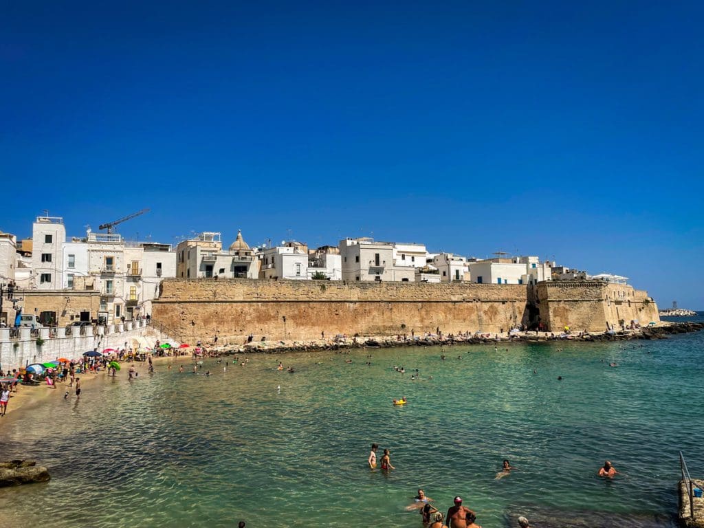 A view of the Old City wall along the ocean in Monopoli, with people swimming at the beach.