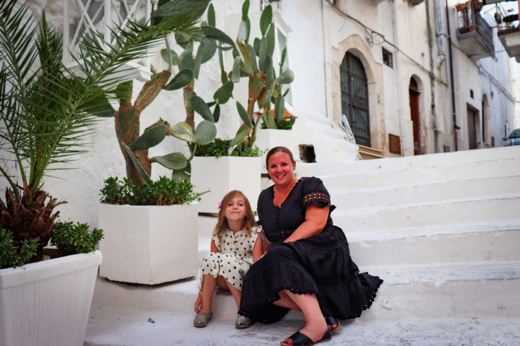 A mom and her young daughter sit together on white-washed steps in Ostuni, one of the best places to visit in Italy on a budget with kids. 