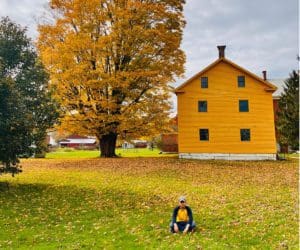 A young boy sits in front of a large yellow building, part of the Hancock Shaker Village in Pittsfield, MA.