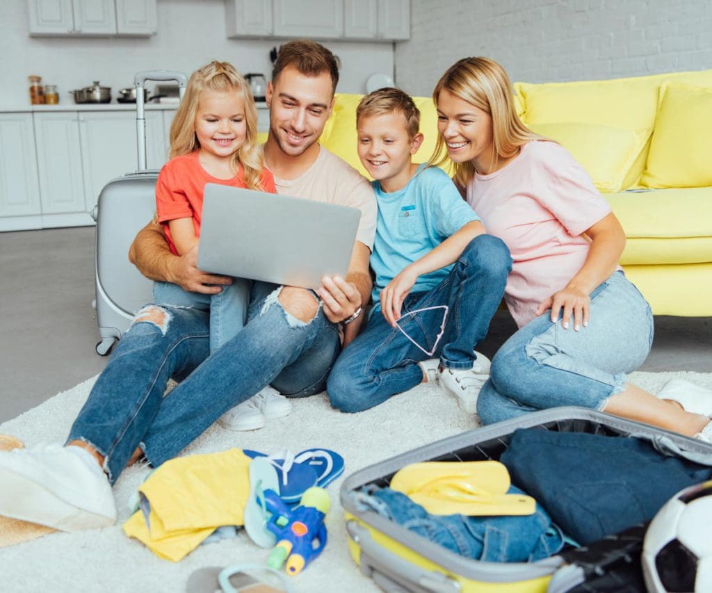 A family of four sits together on the floor, while all looking at a lap top and planning for their next family trip.