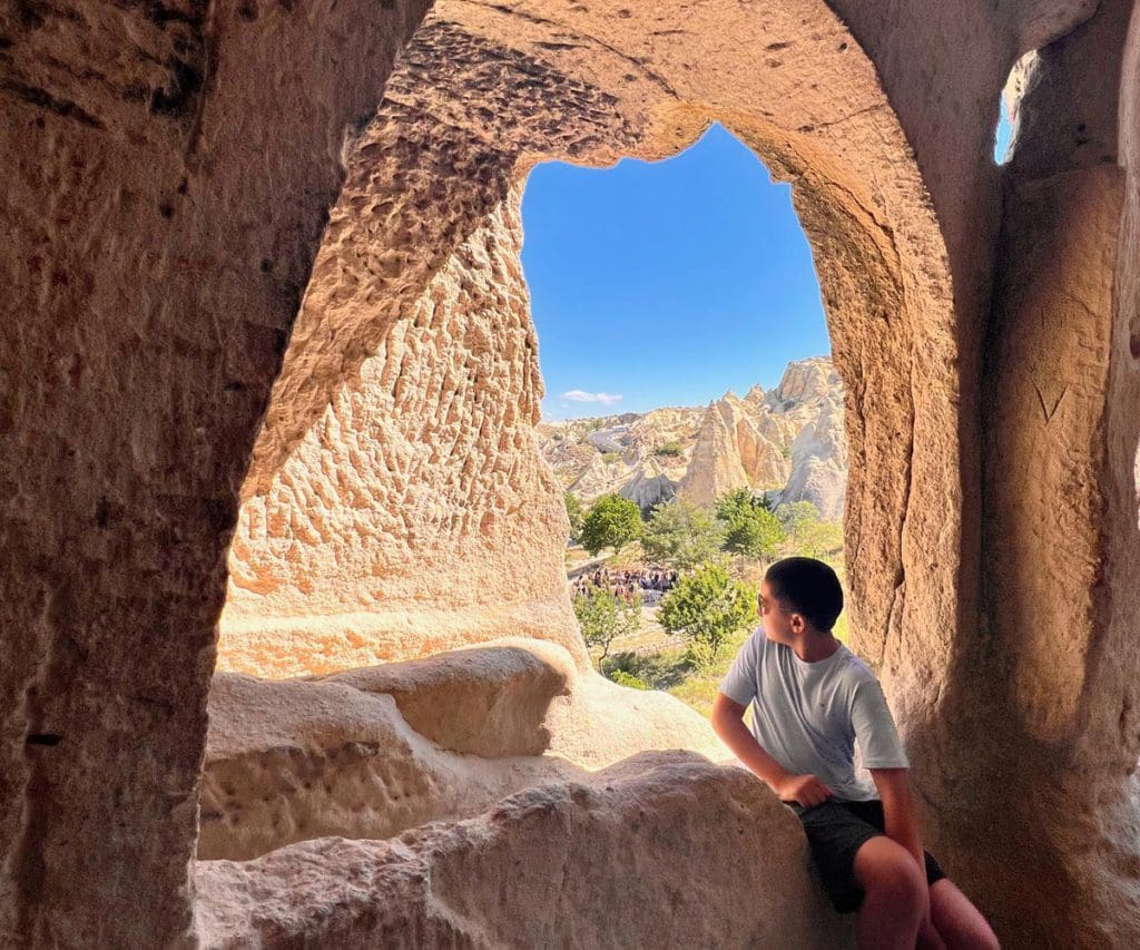 A young boys sits in a cave looking out onto a beautiful view of Cappodocia in Turkey.