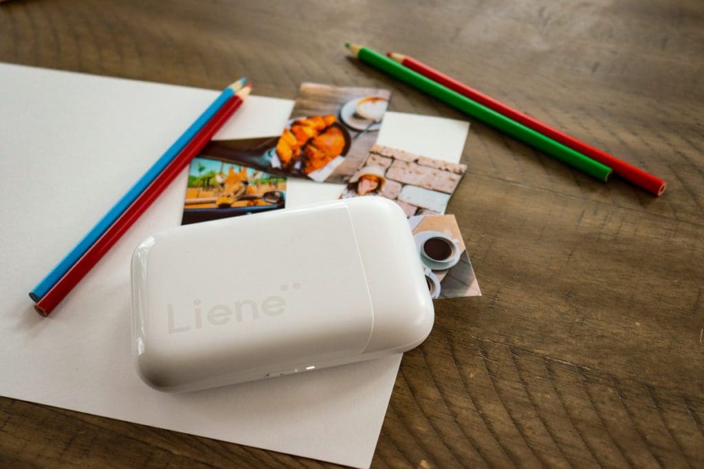 A portable printer prints a travel image, while other lay nearby, as well as paper and colored pencils.