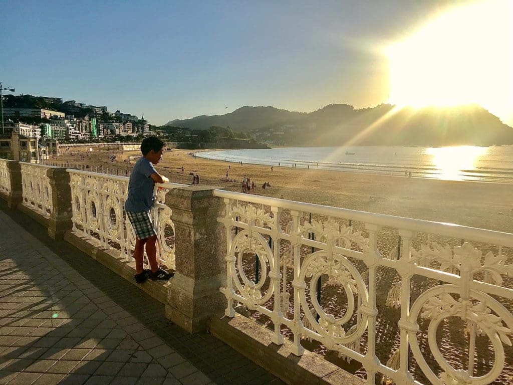 A young boy looks over the railings down to a beach in San Sebastian, Spain at sunset.