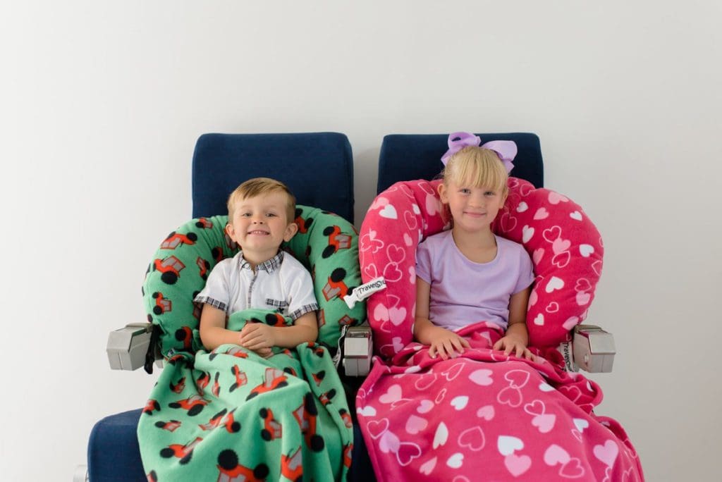 Two kids sit side by side demonstrating their TravelSnugs, one of the best products for sleeping on long international flights with kids.