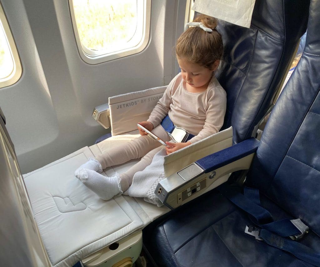 A young girl sits on a plan in her JetKids by Stokke, one of the best products for sleeping on long international flights with kids.