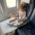 A young girl sits on a plan in her JetKids by Stokke, one of the best products for sleeping on long international flights with kids.