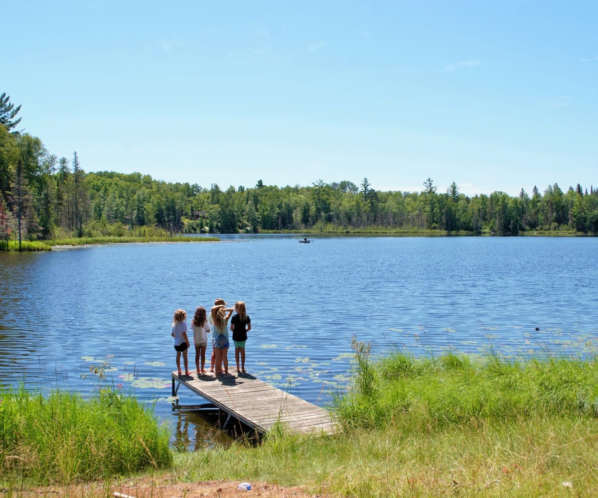 Five kids stand at the end of a dock on a lake in the middle of summer on a sunny day.