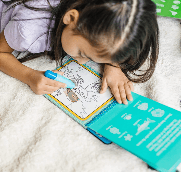 A young girl lays on the floor and colors in a Melissa & Doug Water Wow book.