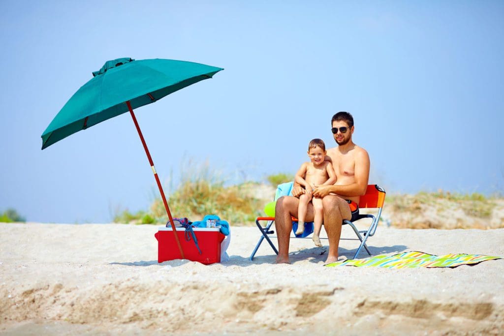A dad and his son sit on a beach chair together near an umbrella and cooler on a sandy beach, a good beach chair is a must on a beach packing list for families.