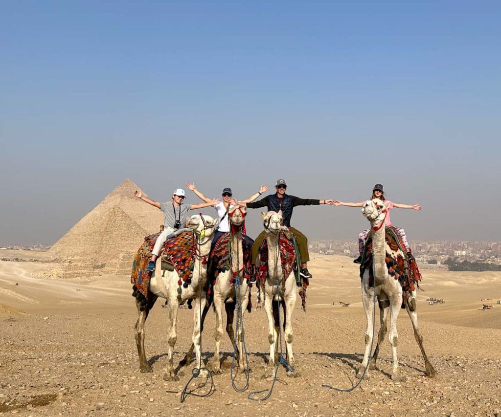A family of four sits atop camels in front of the Pyramids of Giza, a must on this 2-Week Egypt Itinerary for Families.