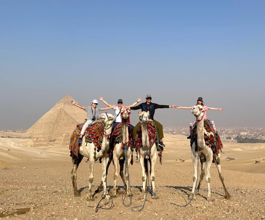 A family of four sits atop camels in front of the Pyramids of Giza, while visiting Egypt with kids.