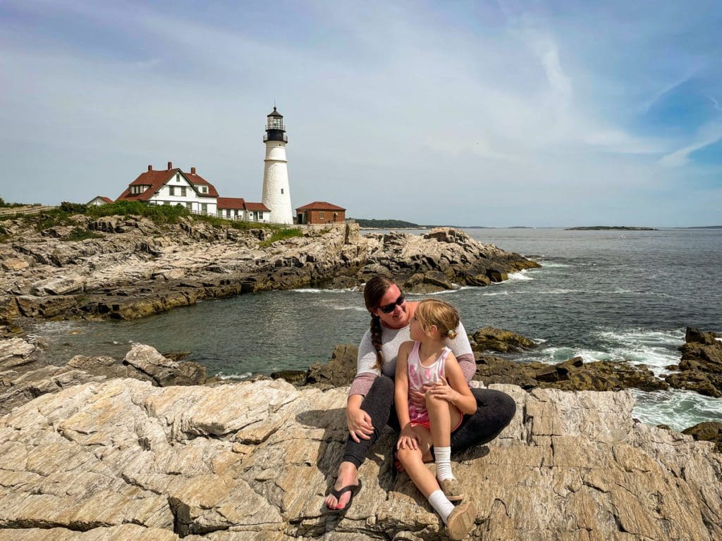 A mom and her young daughter sit together in front of a lighthouse in Cape Elizabeth, Maine.