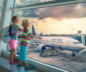 Two kids in brightly colored clothing stand together looking out of a airport window at a United Airlines plane.
