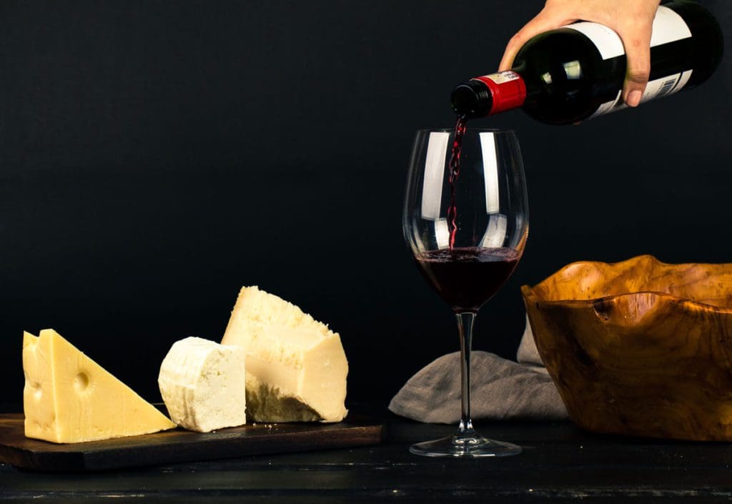 A hand pours a glass of red whine on a table filled with different types of cheese, wine-inspired gifts are amongst the best travel gifts for Mom this Christmas.