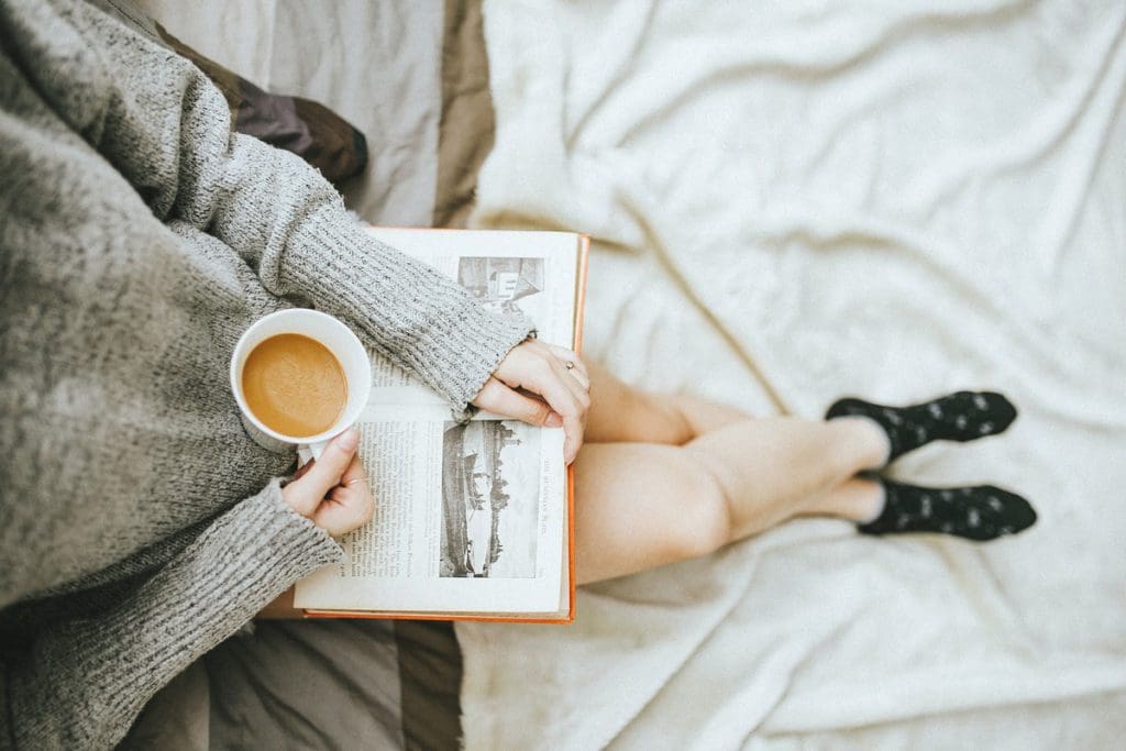 An overhead look down on a woman's lap, where she holds a book and a coffee, and enjoys time alone, one of the best travel gifts for Mothers Day.