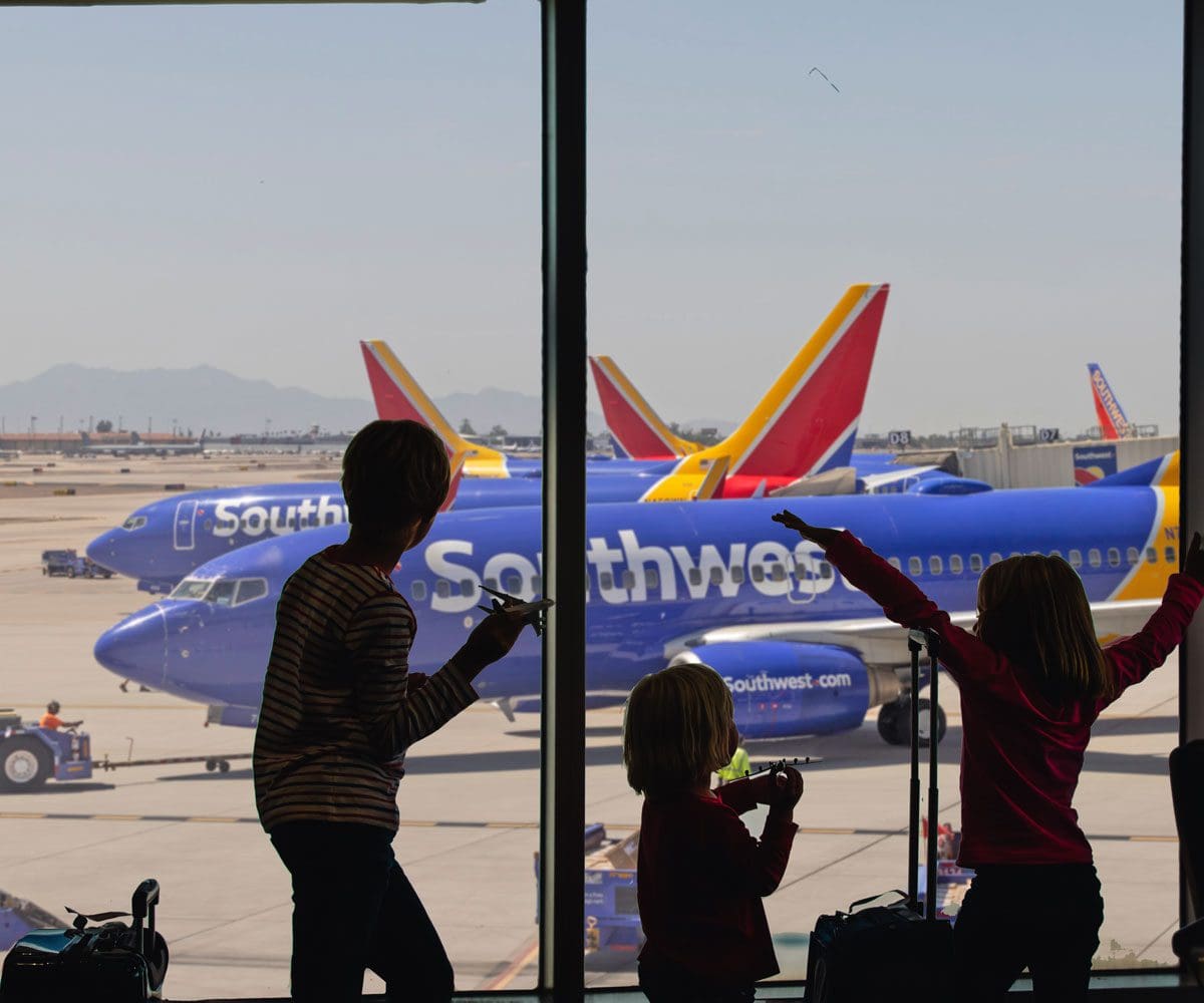 Three kids stand at a window in the airport looking out onto several Southwest planes.