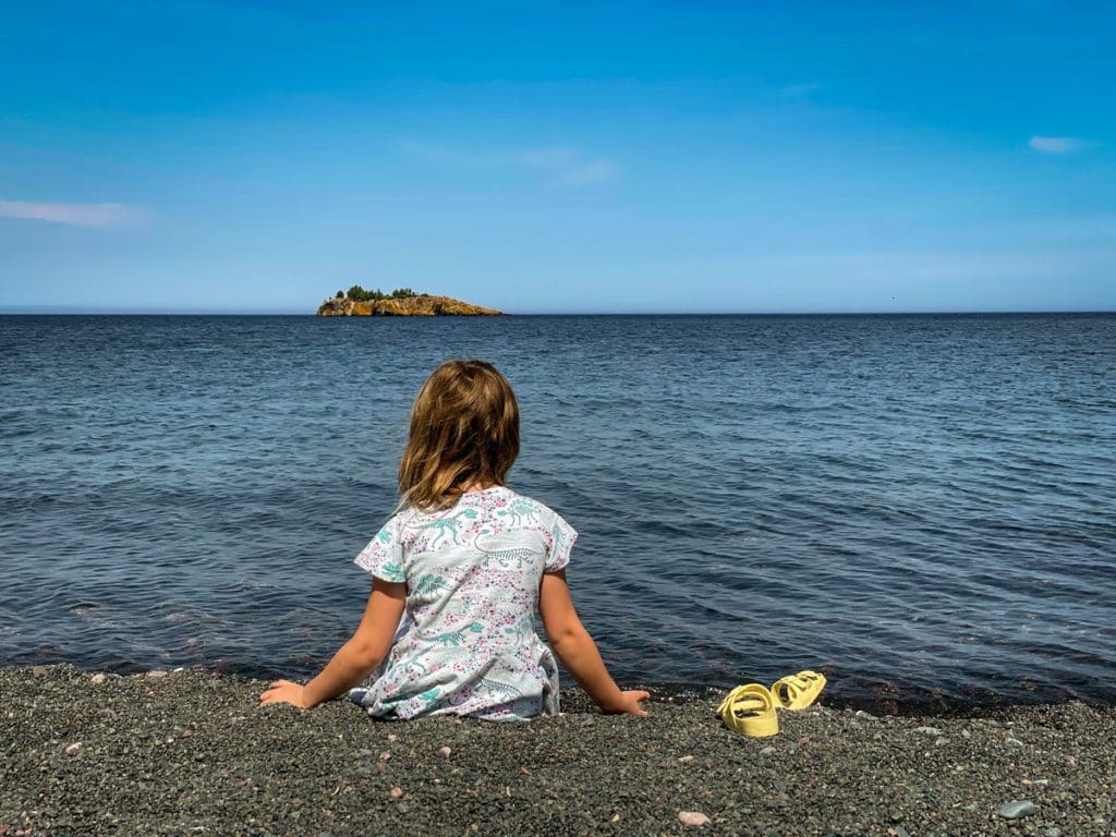 A young girl sits on the shore of Lake Superior looking out toward the water, one of the best lakes in Minnesota for families.