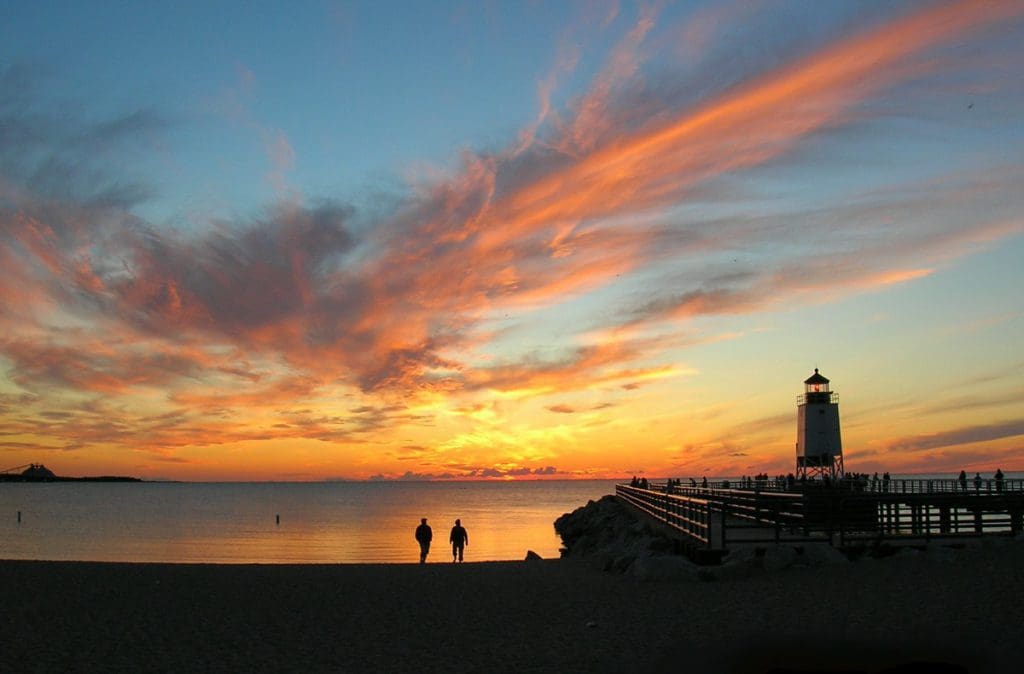 Two people stand silhouetted against a vibrant sunset over Lake Charlevoix.