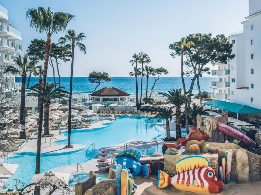 The kid's pool at Iberostar® Alcudia Park, surrounded by palm tress and resort buildings, with a view of the ocean in the distance.