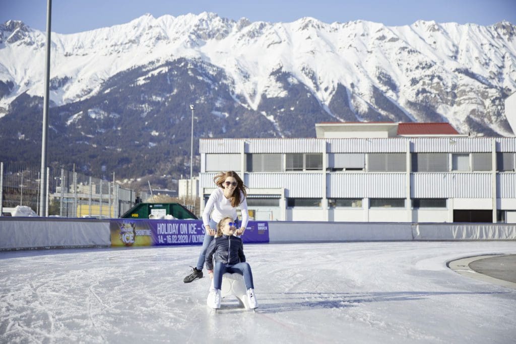 A mom pushes her young daughter on a chair around the ice rink, while skating around, in Innsbruck, one of the best destinations in Austria with kids.
