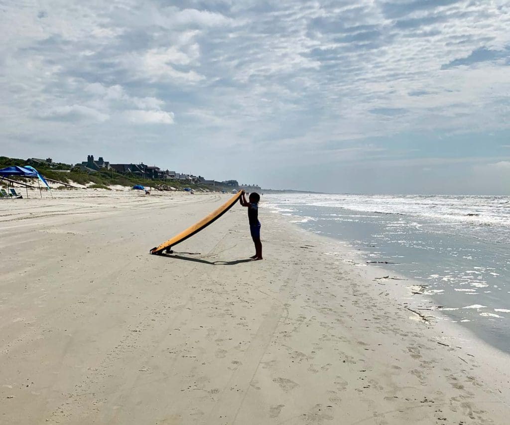A young girl standing on a beach holding a surfboard on Kiawah Island, one of the best beach towns on the East Coast with kids.