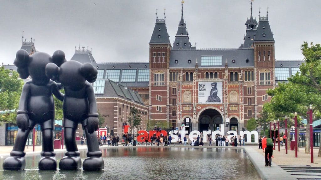 People walk around on a street outside of a prominent Amsterdam building.