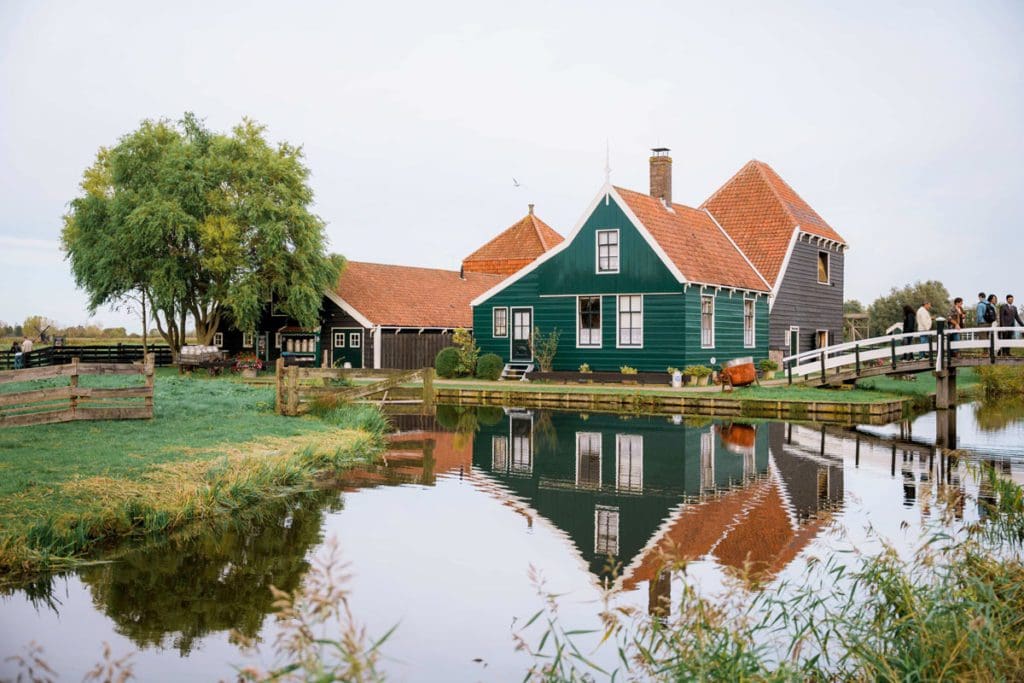 A large green house sits at the edge of a pond in Zaans Schans, with visitors approaching the house by bridge.