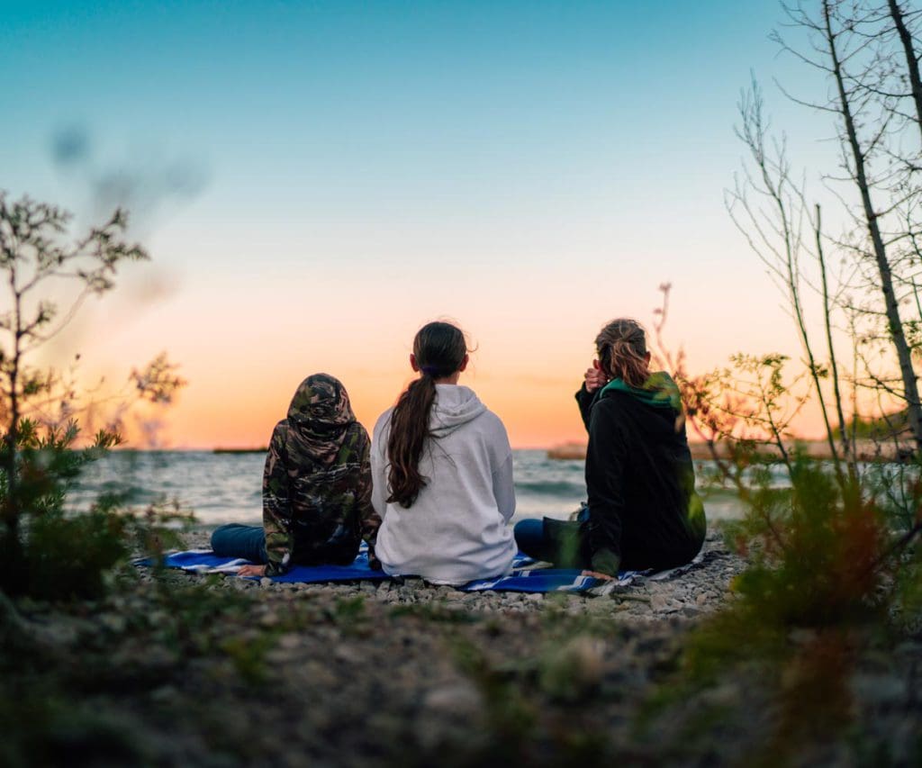 Three kids sit together looking out onto a sunset over Lake Huron.