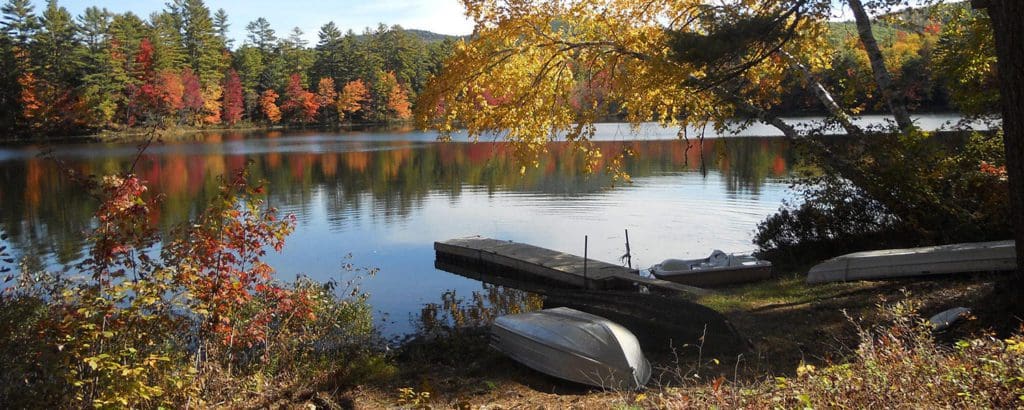 A dock and rowboat rest on the shores of Lake Luzerne during the fall, with colorful trees surrounding the lake.