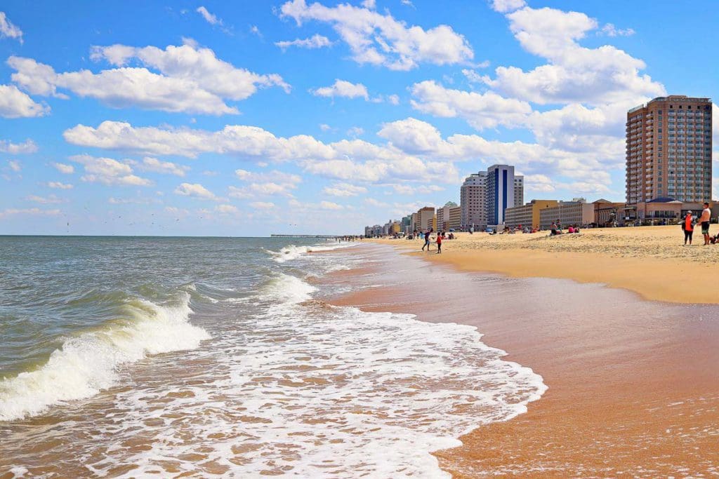 Virginia Beach on a sunny day, a family-friendly beach vacation spot on the East Coast, with people walking on the sandy shore and distant buildings.