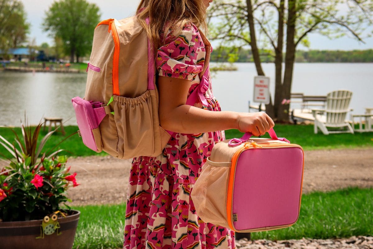 A young girl has a backpack on her back and is carrying a lunch box.