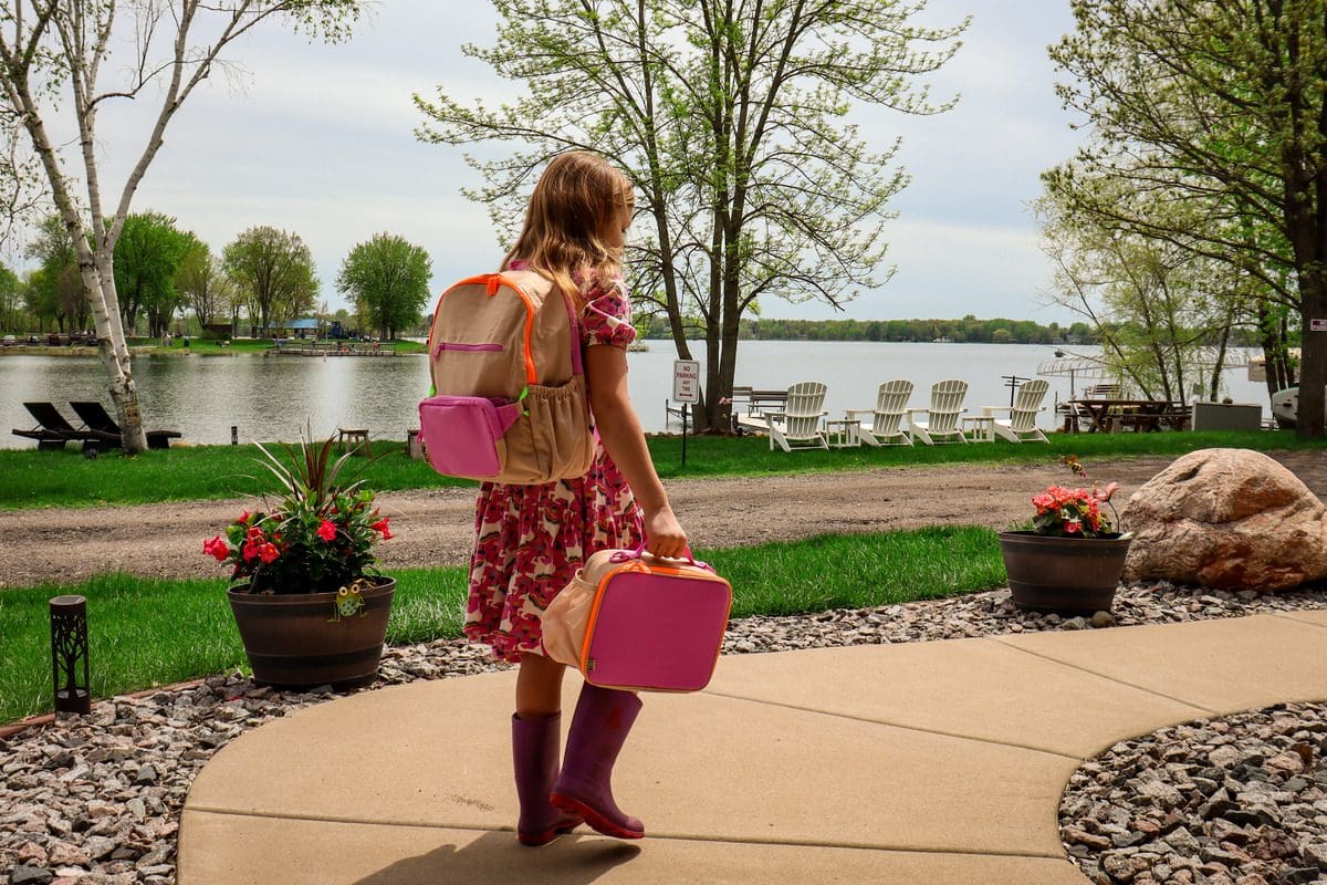 A young girl walks along a sidewalk carrying her backpack and lunch box.