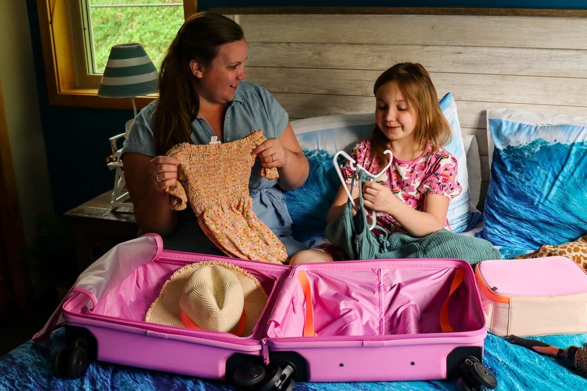 A mom and her young daughter sit on a bed and pack a suitcase together.