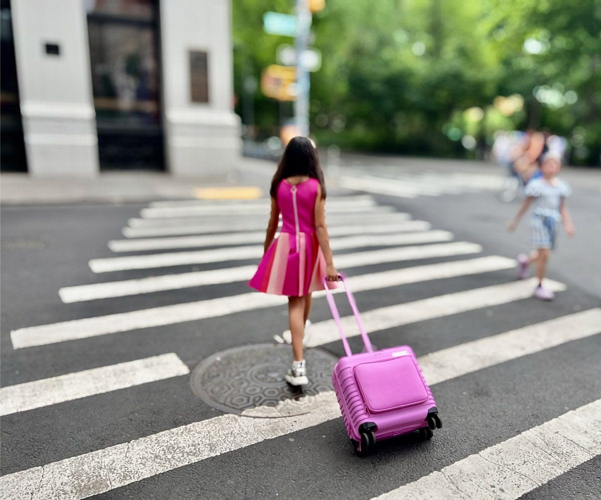 A young girl crosses an NYC street with her pink luggage.