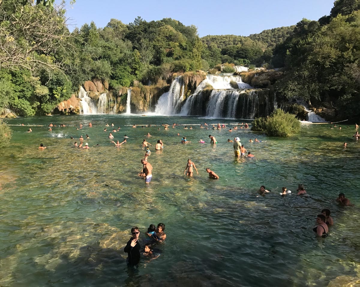 Several people swim in a body of water below a waterfall in Krka National Park, one of the best places to visit in Croatia with kids this summer.