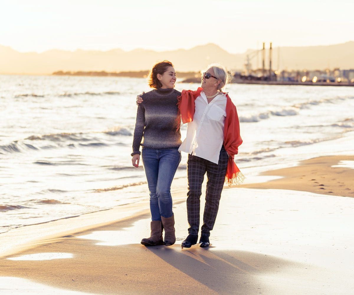 A woman and her elderly mom enjoy a stroll on a beach.
