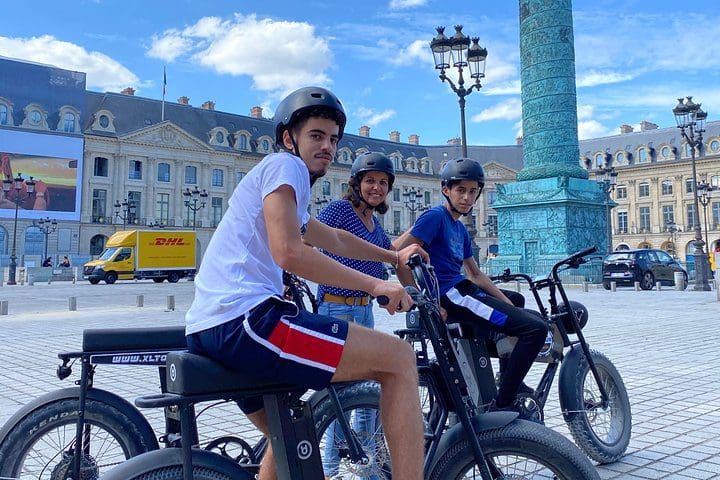 Three kids sit atop their respective electric bikes on a Paris street, while on a Paris Sightseeing Family-Friendly Guided Electric Bike Tour.