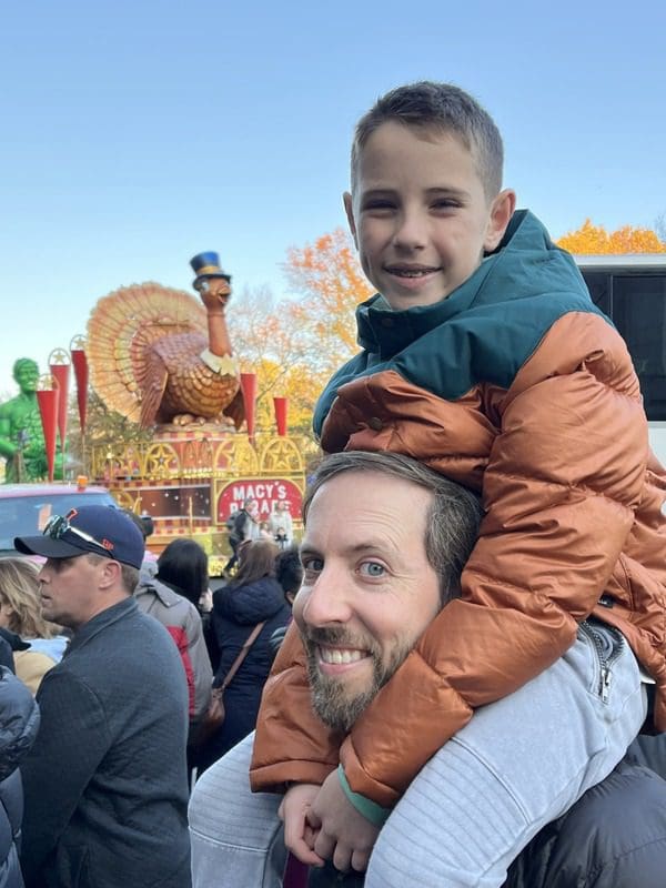 A boy sits atop his dad's shoulders with Turkey Tom in the distance.