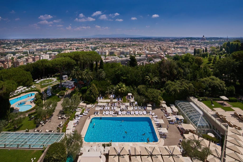 An aerial view of the outdoor pool and grounds at Rome Cavalieri, A Waldorf Astoria Hotel, with a view of Rome.