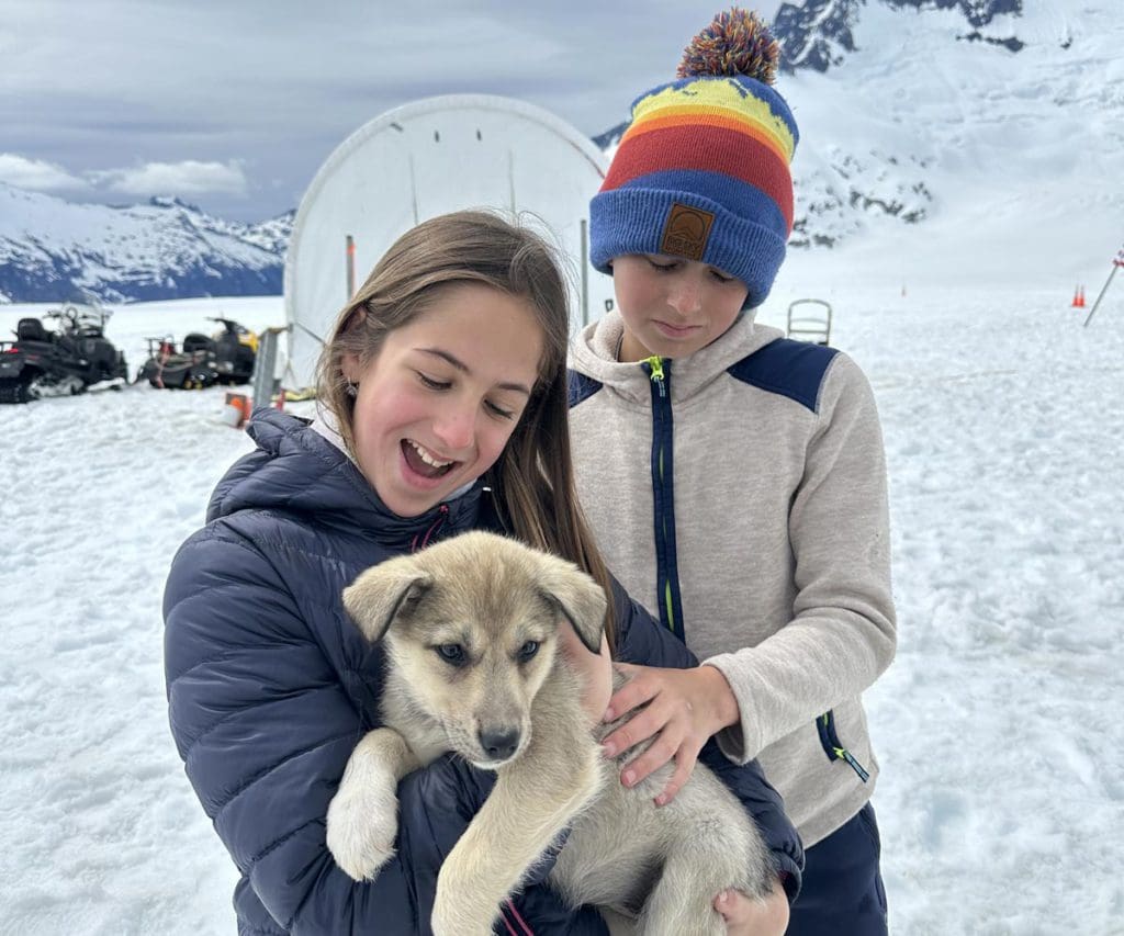 Two kids interact with a husky puppy during a dog sledding experience in Alaska.