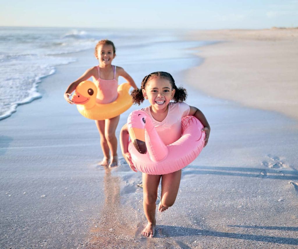 Two girls with large floaties run along a beach, enjoying a warm-weather December vacation.