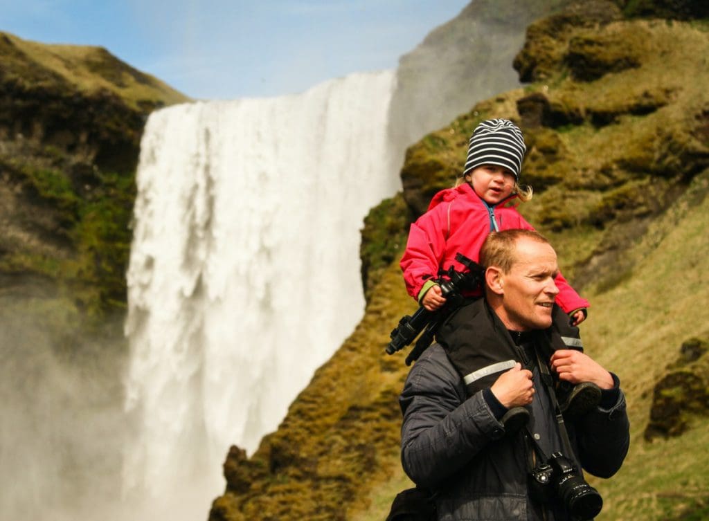 A man holds his young child atop his shoulders while exploring a waterfall in Iceland.