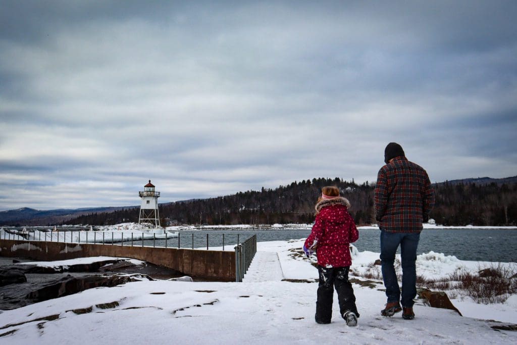 A dad and his young daughter walk through the snow near a light house in Grand Marais, Minnesota, one of the best places for a White Christmas in the United States for families.