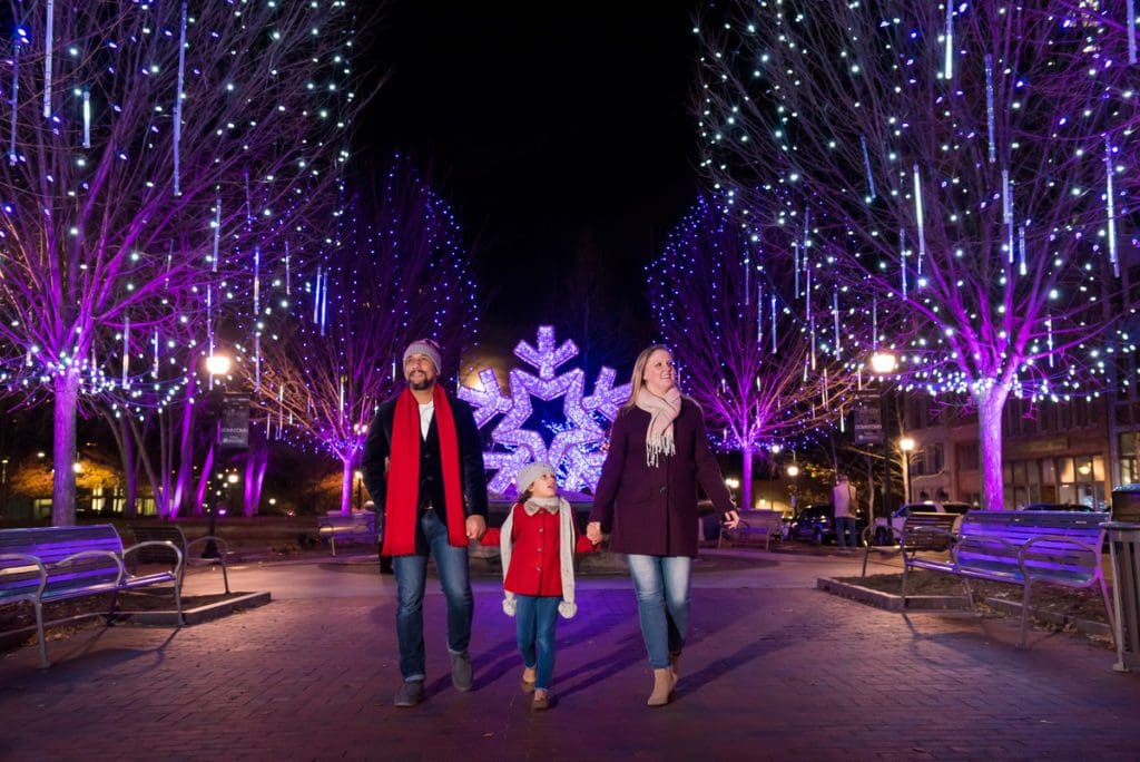 A family of three walks through a path alight with Christmas lights in Asheville, one of the best budget-friendly Christmas destinations in the US for families.