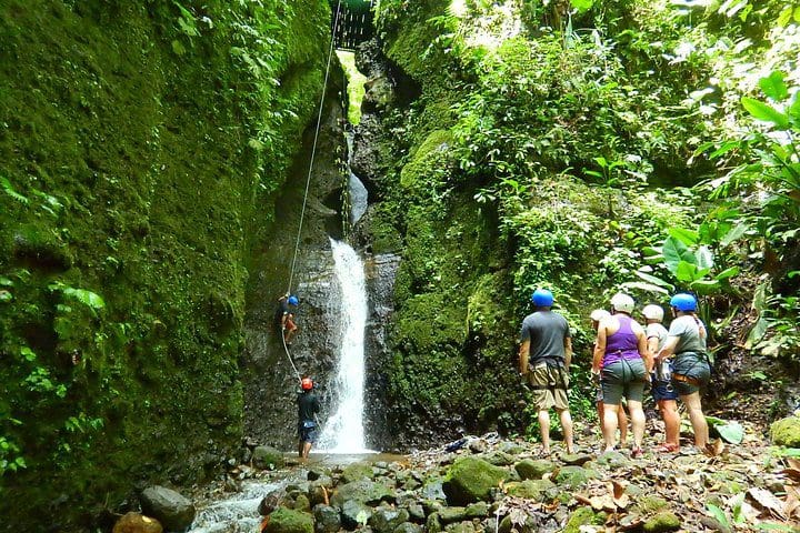 A family stands near a waterfall while experiencing the Pure Trek Canyoning and Waterfall Rappelling Tour in La Fortuna.
