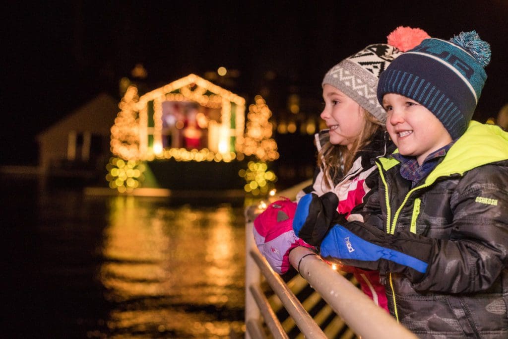 Two kids enjoy a holiday cruise in Coeur d'Alene with holiday lights in the distance.
