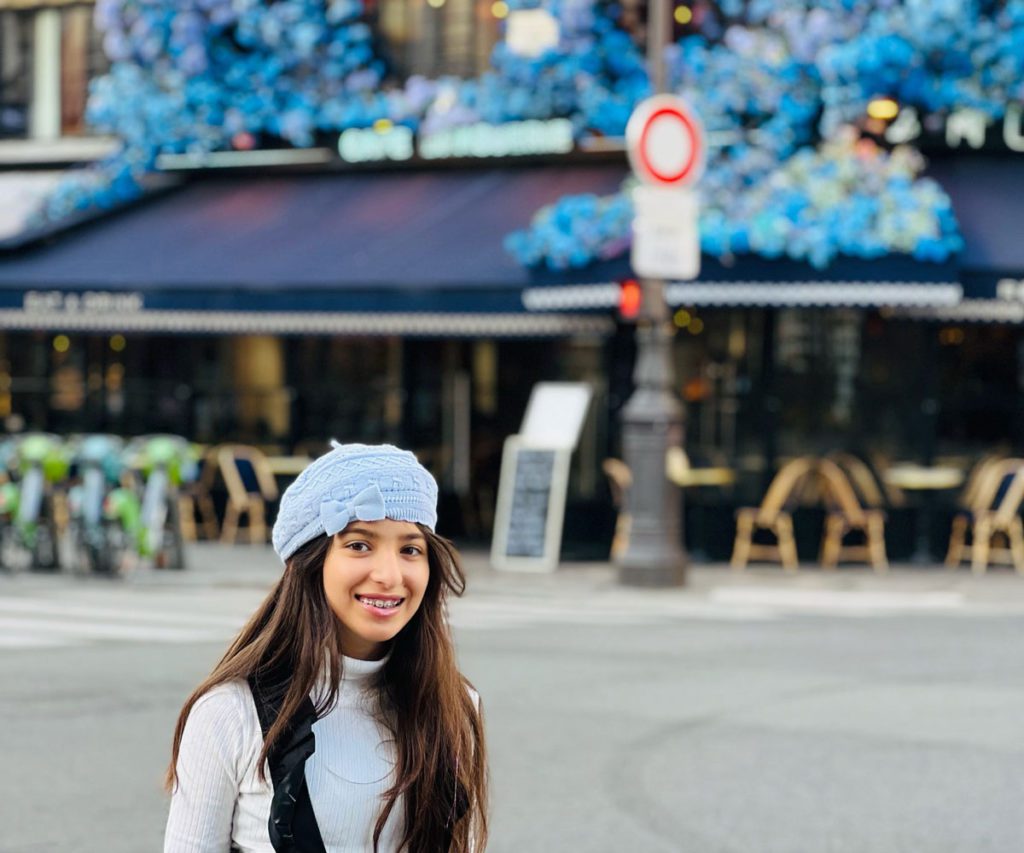 A young girl in a blue hat standing in front of a blue cafe in Paris. 