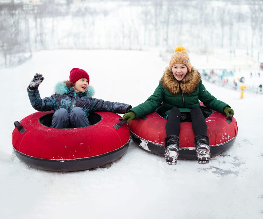 Two kids snow tubing with a red snow tube. 