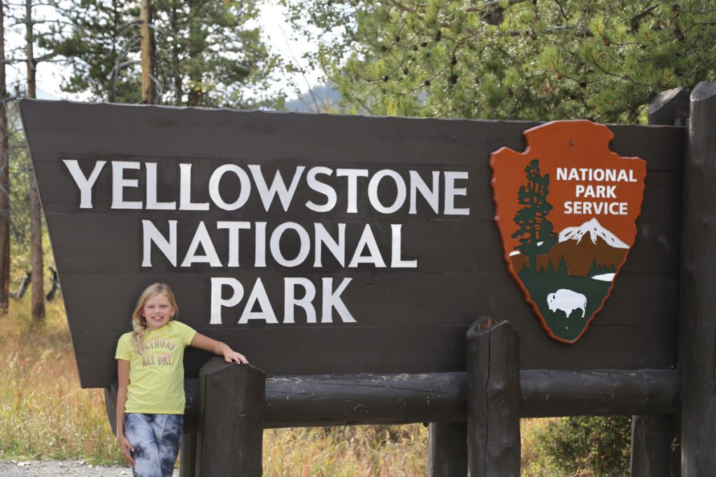 A blonde child standing in front of the Yellowstone National Park sign.