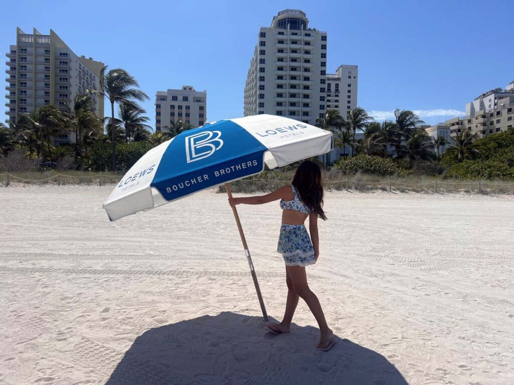 teen girl standing under a Loews Miami Beach-branded umbrella, enjoying the beach with a backdrop of city buildings.